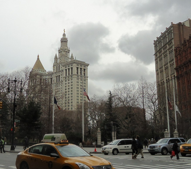 Gelbes Taxi auf einer Stadtstraße mit hohen Gebäuden, Fußgängern auf dem Bürgersteig, Verkehrskegeln, Laternenmasten, Fahnenmasten, Bäumen und einem bewölkten Himmel im Hintergrund.