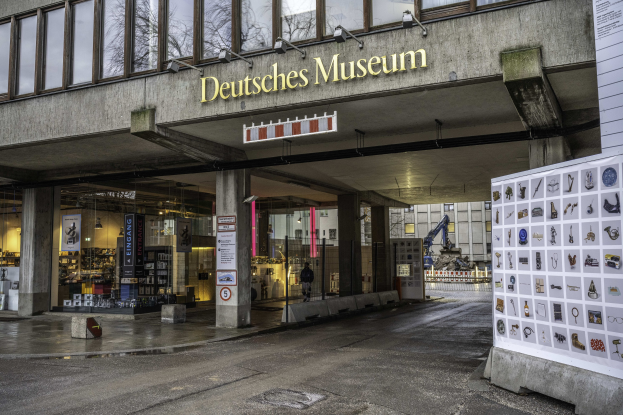 Außenansicht des Deutschen Museums in Berlin, das ein großes Gebäude mit Glasfenstern, Säulen, ein Schild und eine Informationsanzeigetafel auf der rechten Seite sowie andere Gebäude, Bäume und Himmel im Hintergrund zeigt.