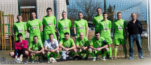 Gruppe von Männern in grünen T-Shirts auf einem Fußballfeld neben einem Torpfosten stehend, mit einem Ball auf dem Boden und verschiedenen Hintergrundelementen wie Bäume, Gebäude, Fahrzeuge.