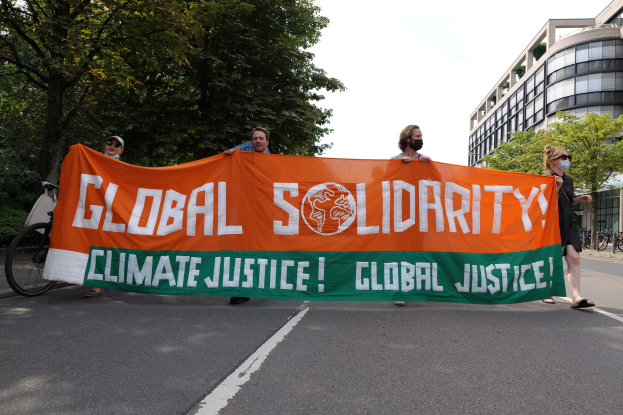 Eine Gruppe von Menschen marschiert eine baumbestandene Straße entlang, die eine "Globale Solidarität: Klimagerechtigkeit"-Tafel hält, mit parkenden Fahrrädern und Gebäuden im Hintergrund.