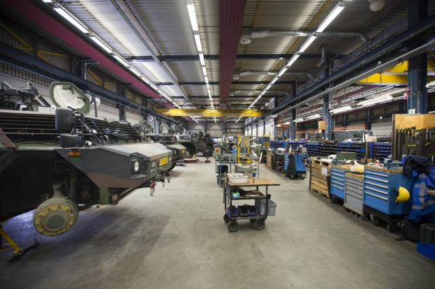 Ein großer Lagerraum mit Maschinen, Fahrzeugen auf der linken Seite, Wagen mit Gegenständen, Holzboxen und anderen Gegenständen auf der rechten Seite und Deckenbeleuchtung.