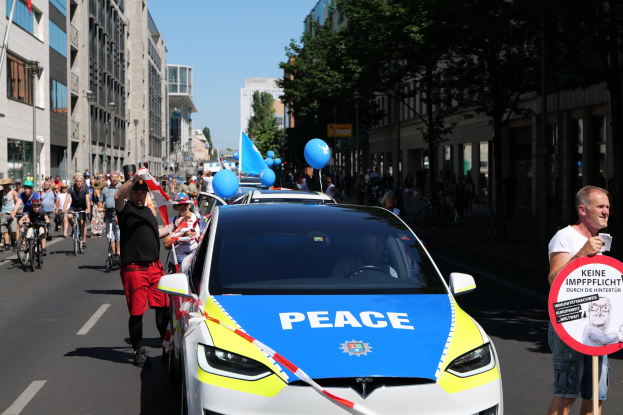 Eine Gruppe von Menschen, die Fahrräder auf einer Straße fahren, mit einem Polizeiwagen im Vordergrund, einer Person, die eine Tafel mit einem Bild und Text hält, und Bäumen, Gebäuden, Laternenmasten und einem klaren blauen Himmel im Hintergrund.