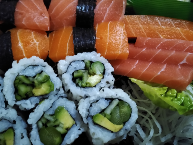 Ein schwarzer Teller mit verschiedenen Sushi-Stücken, darunter Lachs, Avocado und Gurke, umgeben von ein paar grünen Blättern.