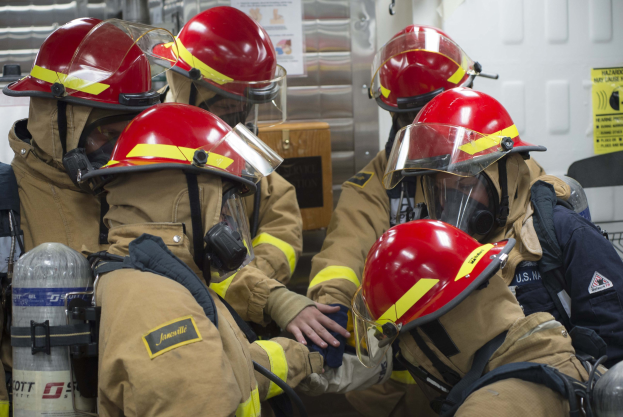 Feuerwehrleute in Schutzausrüstung mit Sauerstoffflaschen löschen ein Feuer, während Plakate und Gegenstände im Hintergrund zu sehen sind.