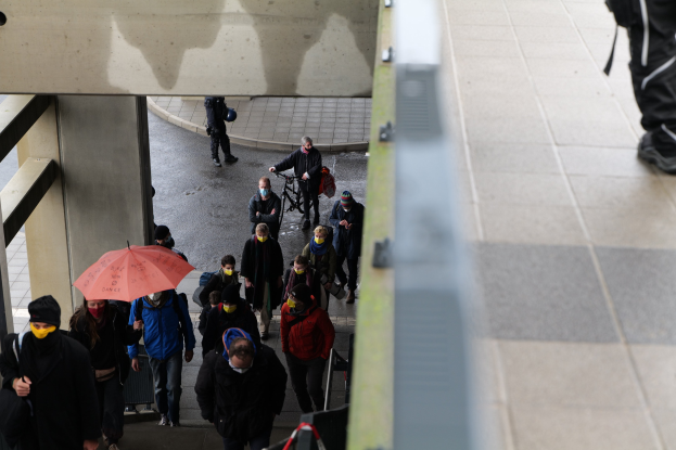 Eine Gruppe von Menschen geht auf einem Gehweg mit Schirmen, einige tragen Taschen und einer hält ein Fahrrad, mit einer Person auf der rechten Seite und Säulen und einer Wand auf der linken Seite, zum Gedenken an ein tragisches Ereignis in Brüssel, Belgien.