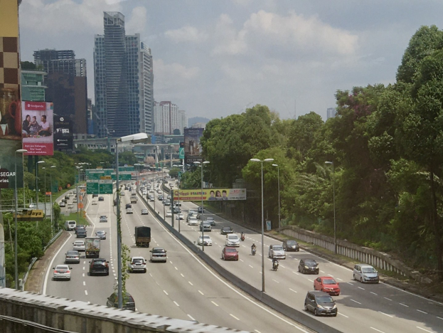 Schwerer Verkehr auf einer Autobahn neben hohen Gebäuden, Straßeninfrastruktur und Bäumen unter einem bewölkten Himmel.