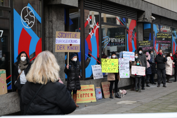 Maskierte Menschen mit Schildern protestieren vor einem Gebäude mit Glaswänden und -türen, auf denen Schilder und Logos zu sehen sind, während Kameras und ein Hund im Bild sind.
