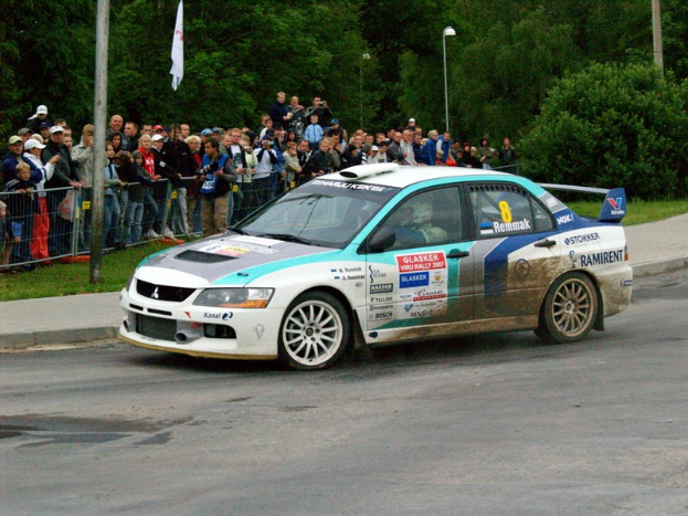 Ein Rallyeauto mit Text und Logos fährt an einer Menge vorbei, die Kameras hält, mit Geländern, Polen mit Fahnen und Bäumen im Hintergrund.