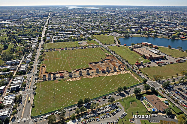 Luftaufnahme einer Stadt mit Gebäuden, Bäumen, Gras, Straßen mit Fahrzeugen, Wasser und Wolken, die einen großen zentralen Platz für einen Park zeigen.
