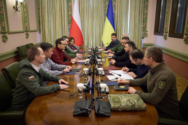 Gruppe von Männern in formeller Kleidung, die um einen Holztisch mit Mikrofonen und Dokumenten sitzen und in einem Raum mit Flaggen, Vorhängen und an der Wand montierten Lampen diskutieren.