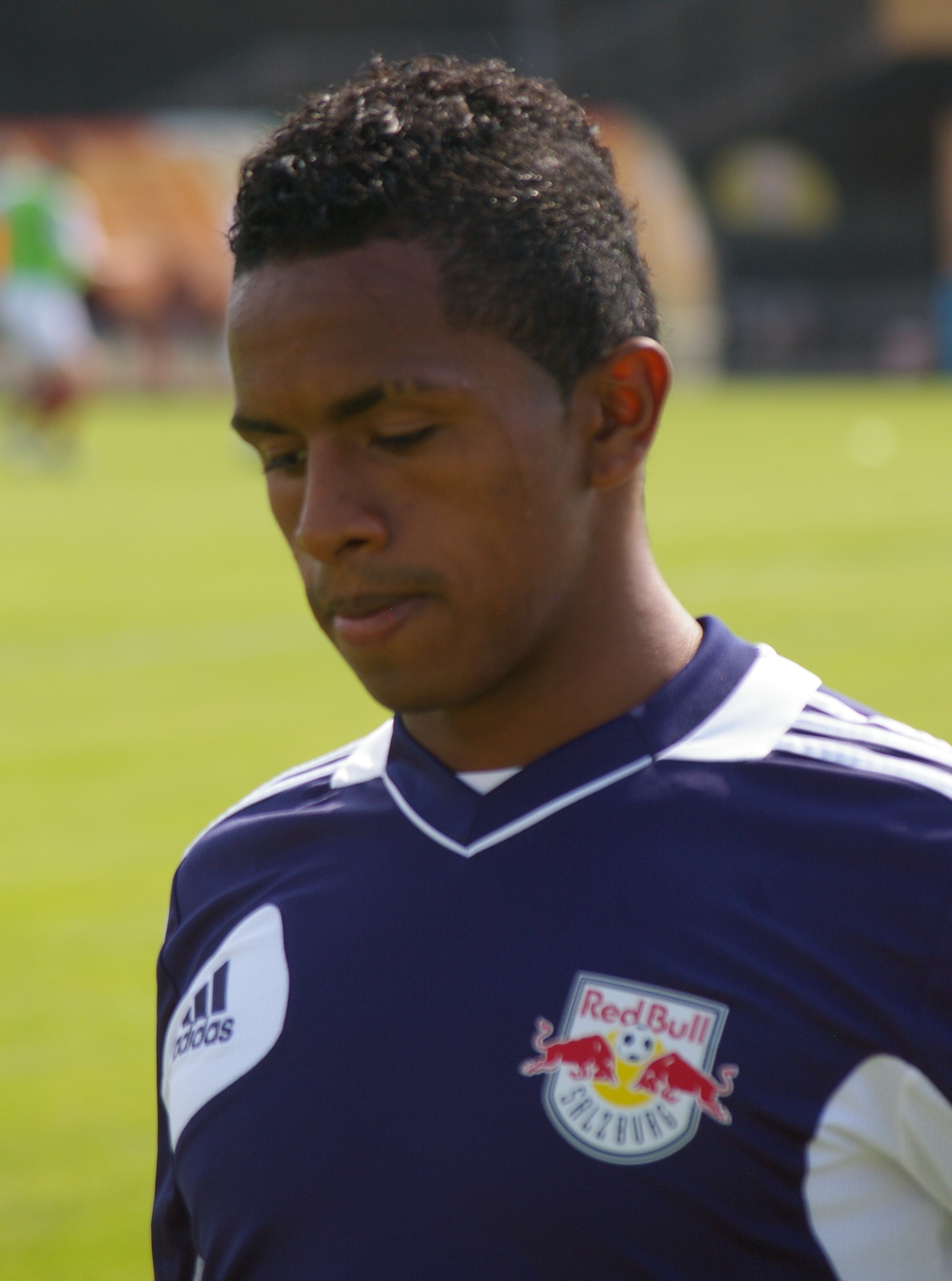 Ein junger Mann in einem blauen T-Shirt steht auf einem Fußballfeld, mit kurzen dunklen Haaren und einem entschlossenen Gesichtsausdruck, der als Flügelspieler mit einem unscharfen Hintergrund erscheint.
