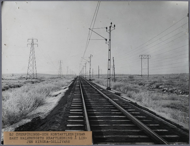 Schwarzes und weißes Foto einer Schienenstrecke mit elektrischen Masten und Drähten, die daneben verlaufen, umgeben von Gras auf beiden Seiten, mit Text unten.