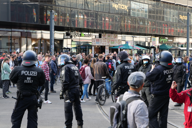Eine Gruppe von Polizisten vor einer Menschenmenge auf der Straße, einige halten Fahrráder und andere tragen Helme und Taschen, vor dem Hintergrund von Gebäuden mit Glaswänden, Namensschildern, Laternenmästen und Schirmen.