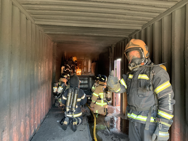 Feuerwehrleute in Schutzausrüstung, einschließlich Helmen, Handschuhen und Sauerstoffflaschen, arbeiten innerhalb eines Seecontainers mit einem Feuerlöscher im Hintergrund.