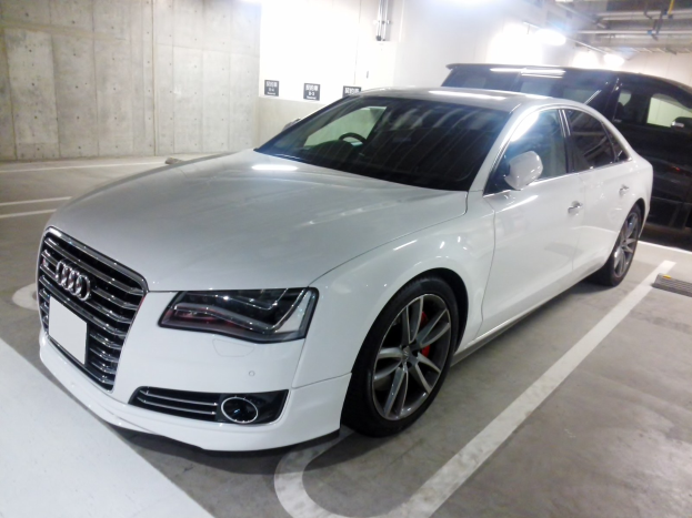 Ein weißer Audi S8 in einer Tiefgarage geparkt neben einem schwarzen Auto, mit sichtbaren Garagenwänden, Deckenrohren und Lampen.