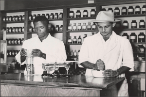Zwei Männer in Laborkitteln und Hüten stehen an einem Tresen in einer Apotheke mit einer Waage und einer Schüssel, mit Regalen voller Flaschen im Hintergrund, dargestellt in Schwarz-Weiß.
