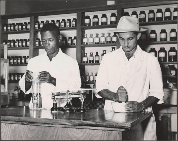 Zwei Männer in Laborkitteln und Hüten stehen an einem Tresen in einer Apotheke mit einer Waage und einer Schüssel, mit Regalen voller Flaschen im Hintergrund, dargestellt in Schwarz-Weiß.