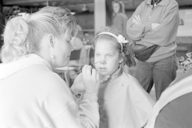 Ein junges Mädchen lässt sich von einer Frau in einem Salon Make-up auflegen, während zwei andere Personen im Hintergrund stehen, alles in Schwarz-Weiß.