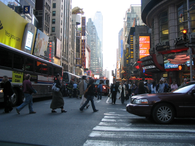 Eine belebte Stadtstraße mit Fahrzeugen, Fußgängern an einer Zebrastreifenfurt, Gebäuden, Schildern, Verkehrsampeln und einem klaren blauen Himmel.