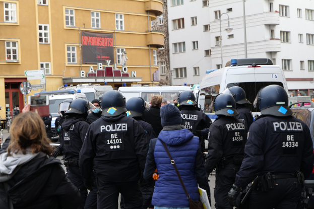 Eine Gruppe von Polizisten in Uniform steht vor einer Menge von Helm- und Jackentragenden Protestierenden, mit Fahrzeugen, Gebäuden, Laternenmästen und einem Banner im Hintergrund, während eine Person mit einer Kamera die Szenerie dokumentiert.