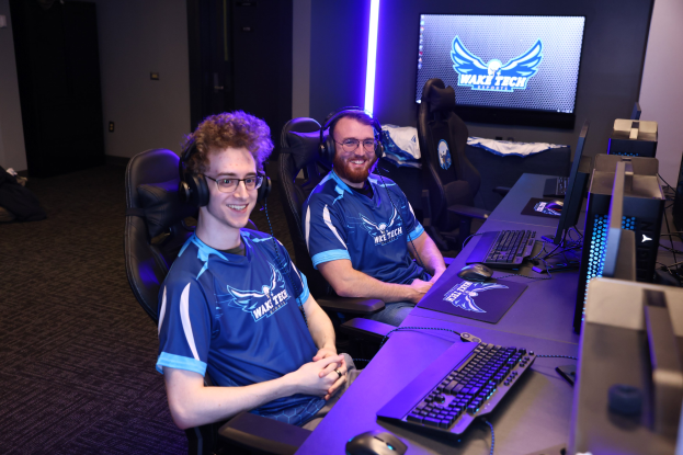 Zwei Männer mit Headsets sitzen an einem Schreibtisch mit Monitoren, Tastaturen und anderer Ausrüstung und lächeln, während sie ein Spiel von Wake Tech League of Legends spielen.