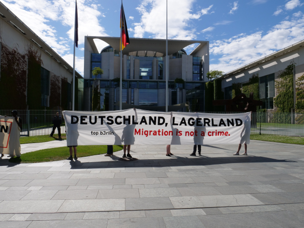 Gruppe von Menschen, die eine Fahne mit der Aufschrift "Deutschland, Lagerland Migration ist kein Verbrechen" tragen, vor einem Gebäude mit Fenstern, umgeben von Gras, einem Metallzaun, Pflanzen, Bäumen und Fahnenmästen.