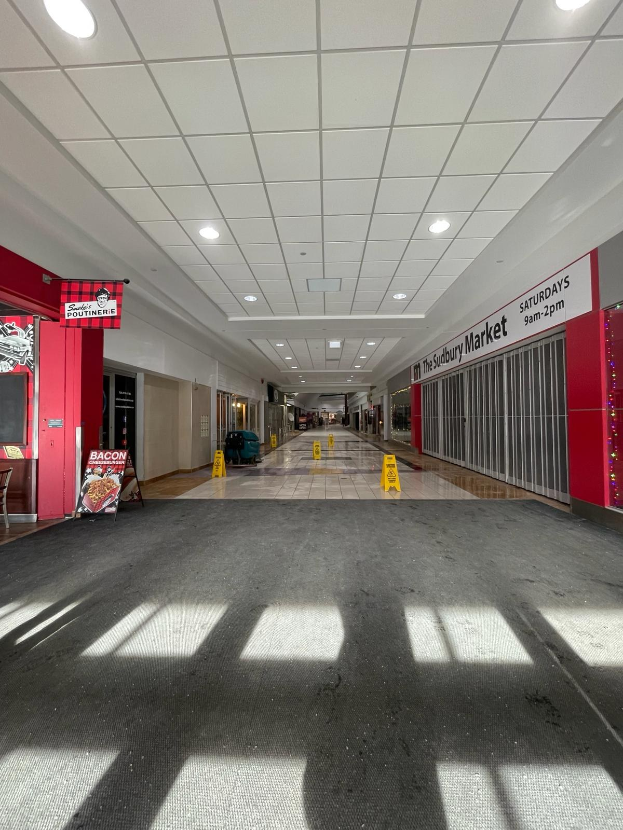 Innenansicht eines Einkaufszentrums mit Deckenbeleuchtung, einem Stuhl und einer Tafel, auf der "Sydbury Market - Vorher und Nachher" steht, auf der linken Seite und verstreuten Gegenständen auf der rechten und auf dem Boden.