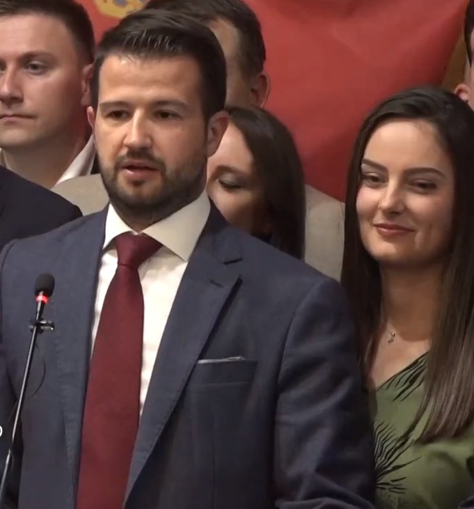 Ein Mann im Anzug spricht in ein Mikrofon an einem Rednerpult, mit einem Banner im Hintergrund und einer sichtbaren Menge, wahrscheinlich spricht er über ein Regierungsverbot der sozialen Medien.