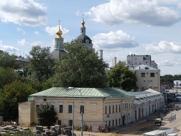 Eine Stadtansicht mit der Kirche der Interzession an der Nerl im Hintergrund, umgeben von Gebäuden, Bäumen, Straßeninfrastruktur, Fahrzeugen, Fußgängern und einer bewölkten Himmel.