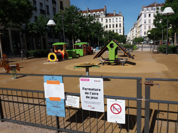 Ein umzäunter Stadtplatz-Spielplatz mit mehreren Schildern an der Umzäunung, umgeben von Bäumen, Laternen, Gebäuden mit Fenstern und einem sichtbaren Himmel, mit verstreuten Spielplatz-Objekten.