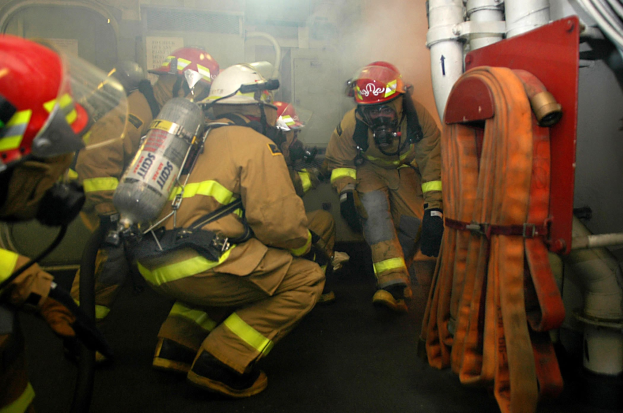 Feuerwehrleute in Schutzausrüstung mit Rauch aus dem Mund, die in einem raucherfüllten Raum mit Rohren und einer Wand im Hintergrund stehen.