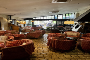 Eine geräumige Hotellobby mit zahlreichen Stühlen und Tischen, gepolsterten Sofas, an der Wand angebrachten Lampen, einer Deckenklimaanlage und großen Glasfenstern an der Rückseite.