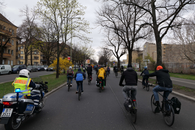 Eine Gruppe von Menschen auf Fahrrädern, die eine von Bäumen gesäumte Straße mit Gebäuden auf beiden Seiten entlangfahren, begleitet von einem Polizeimotorradfahrer auf der linken Seite.