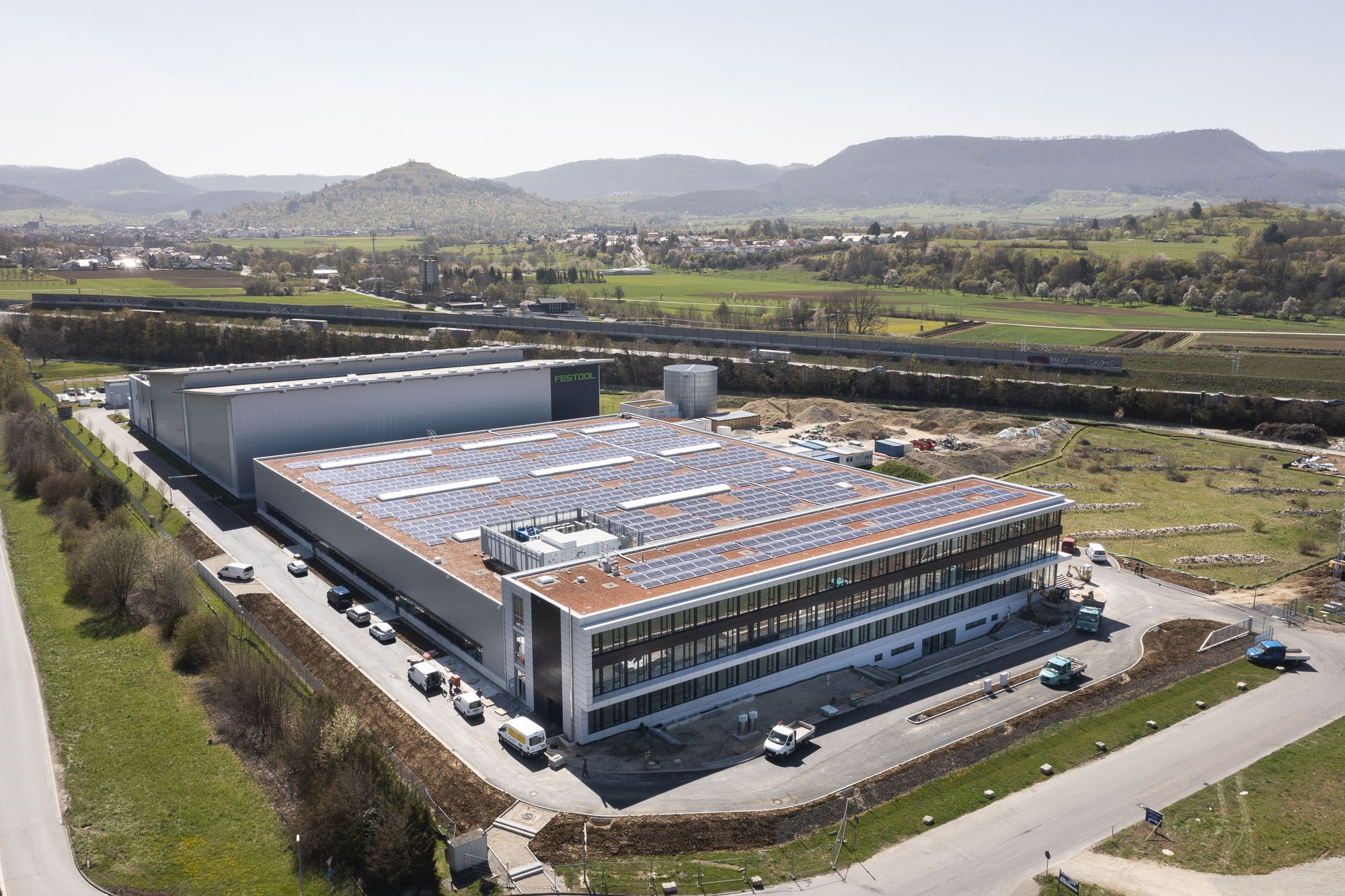 Luftaufnahme eines großen Gebäudes mit Solarmodulen auf dem Dach, umgeben von Fahrzeugen, Bäumen, Gras und Masten, vor den Hügeln und einem klaren blauen Himmel - die erste solarbetriebene Fabrik Deutschlands.