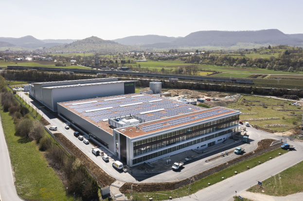 Luftaufnahme eines großen Gebäudes mit Solarmodulen auf dem Dach, umgeben von Fahrzeugen, Bäumen, Gras und Masten, vor den Hügeln und einem klaren blauen Himmel - die erste solarbetriebene Fabrik Deutschlands.