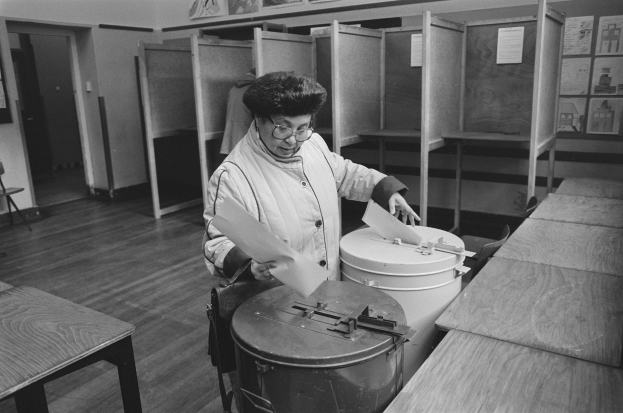 Eine Frau mit Brille hält Papiere in der Hand, während sie in einer Wahllokal abstimmt, mit Tischen, Bänken, Schränken, einer Tür, einem Stuhl und Plakaten im schwarz-weißen Bild.