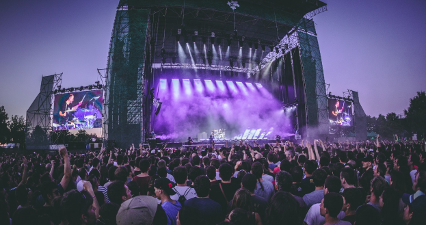 Eine große Menschenmenge bei einem Musikfestival schaut auf einer hell erleuchteten Bühne mit Bildschirmen, die vor einem Gebäude, Bäumen und einem klaren blauen Himmel aufgebaut sind.