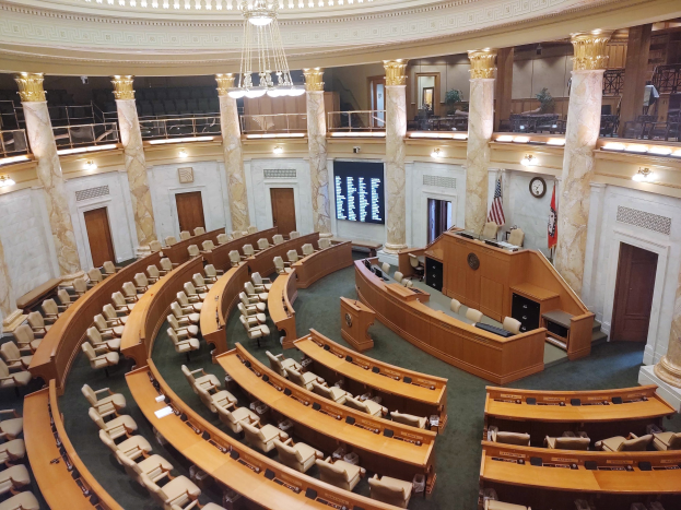 Großer Gesetzgebungsausschuss mit Reihen von Tischen und Stühlen, einem zentralen Podium mit Mikrofon, Flaggen, einer Wanduhr, Türen, Geländern, Deckenlampen und Kronleuchtern.