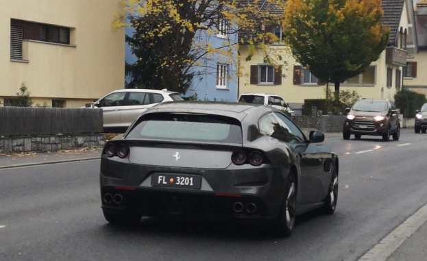 Roter Ferrari GTC4Lusso fährt auf einer Straße mit Gebäuden, Bäumen und einem Zaun im Hintergrund unter einem klaren Himmel.
