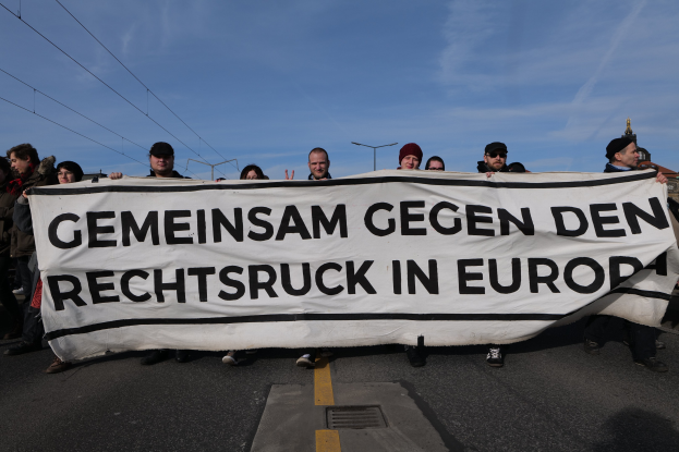 Eine Gruppe von Menschen auf der Straße mit einem Banner, auf dem "Gemeinsam gegen den Rechtsruck in Europa" steht, vor Straßenlaternen, Strommasten, Stromkabeln, Gebäuden und einem bewölkten Himmel.