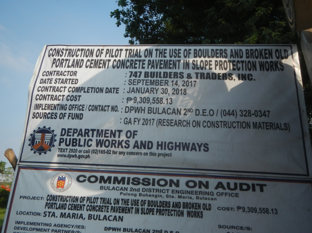 Straßenschild, das einen Pilotversuch zur Verwendung von Steinen und recycelten Portland-Zement-Beton im Hangschutz anzeigt, mit Grünfläche und einem klaren Himmel im Hintergrund.