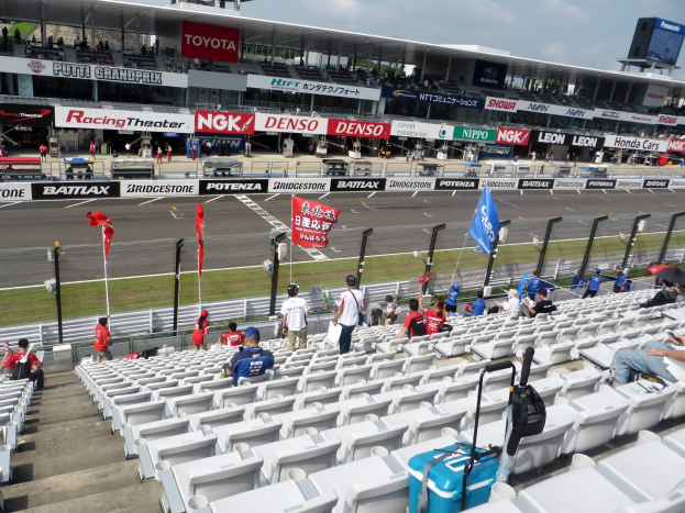Eine Gruppe von Menschen auf Tribünen bei einer Rennstrecke, umgeben von Fahnen, Texttafeln, Geländern und Stufen, mit einem Stadion und einem bewölkten Himmel im Hintergrund.