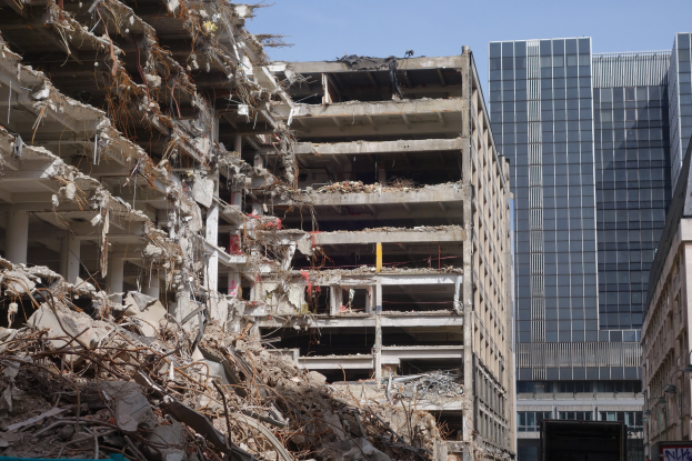Ein großes Gebäude wird in der Mitte einer Stadt abgerissen, umgeben von intakten Strukturen, Straßeninfrastruktur, Fahrzeugen, Bäumen und einem klaren Himmel.