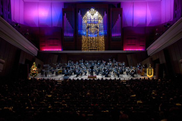 Großer Saal mit Publikum bei einem Konzert, Musiker auf der Bühne mit Instrumenten und sitzende Musiker, Weihnachtsbaum und geschmückte Wände im Hintergrund.