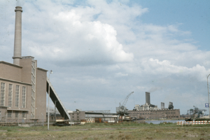 Ein altes Foto einer Fabrik mit Rauch, der aus ihren Schornsteinen aufsteigt, umgeben von Gebäuden, Kränen, Polen, Lampen und Schildern.