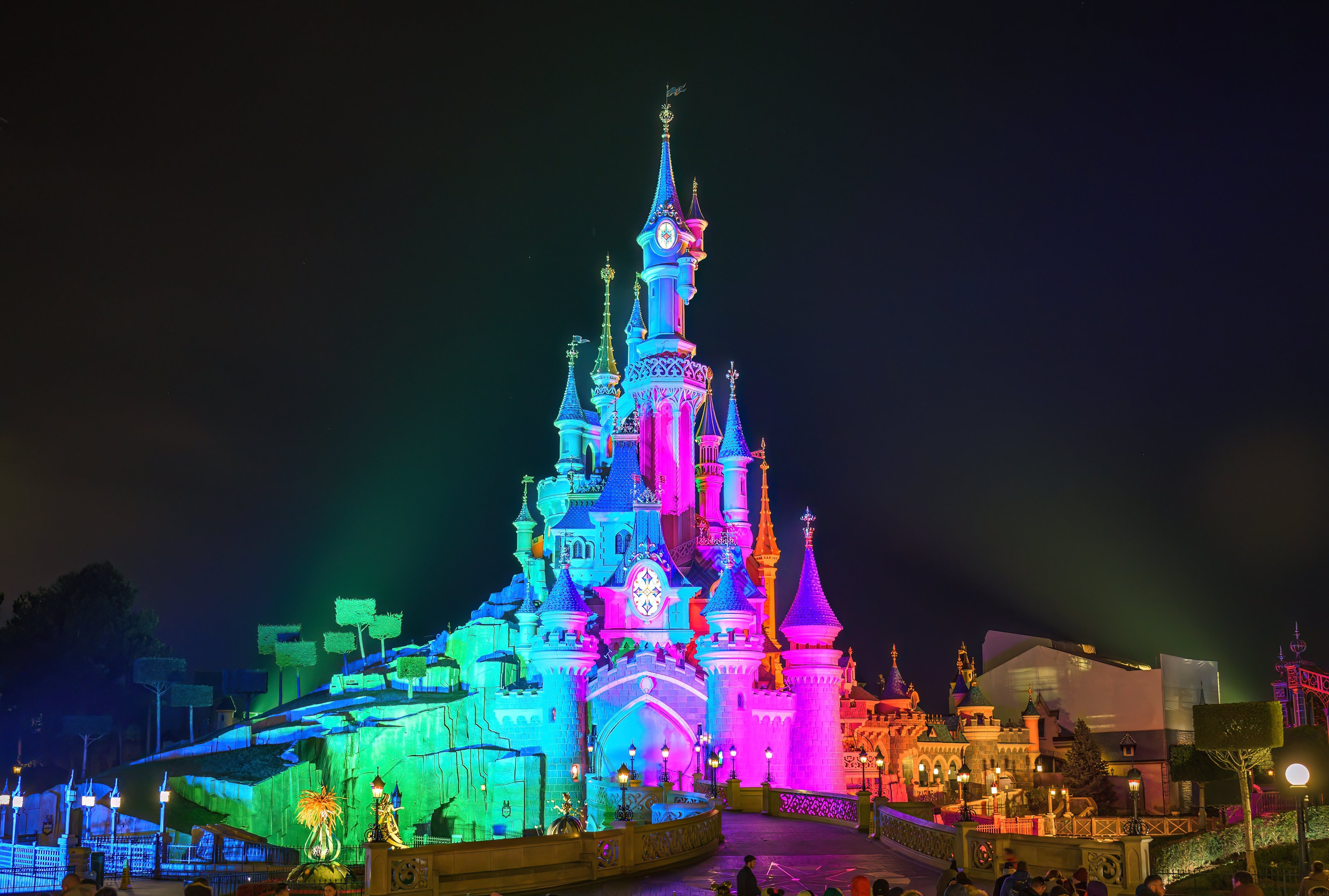 Schloss der Schlafenden Schönheit im Disneyland Paris bei Nacht, hell erleuchtet mit bunten Lichtern, umgeben von Menschen, einem Zaun, Straßenlaternen, Lichtern, Bäumen und Gebäuden unter einem bewölkten Himmel.