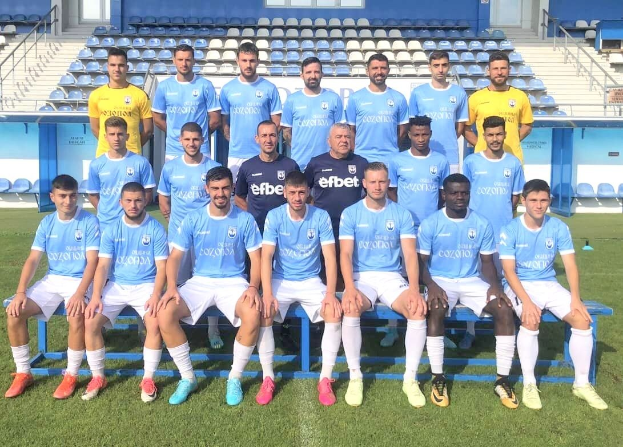 Eine Gruppe von Männern, einige sitzen auf Bänken und andere stehen auf dem Rasen, auf einem Fußballfeld mit Tribünen und architektonischen Elementen im Hintergrund.