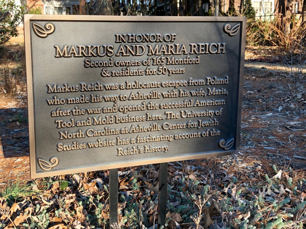 Gedenktafel für Markus und Maria Reich vor einem Gebäude angebracht, umgeben von Bäumen und Pflanzen, mit trockenen Blättern auf dem Boden.