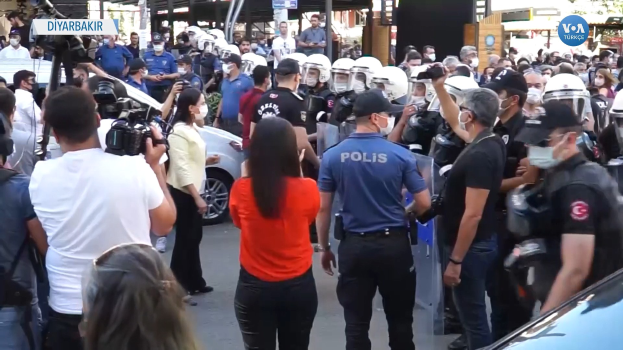 Eine Gruppe von Menschen vor einer Reihe von Polizisten in Helmen, einige mit Kameras, mit Fahrzeugen, Gebäuden, Bäumen und Schildern mit der Aufschrift "türkische Polizei im Konflikt mit Demonstranten in Ankara" im Hintergrund.