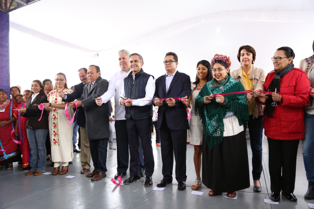Eine Gruppe von Menschen hält eine Schere bei einer Eröffnungszeremonie vor einer Menge, mit einer Säule links, verstreuten Papieren auf dem Boden und einem weißen Tuch im Hintergrund.